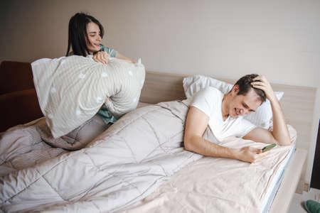 Couple spending weekend morning together in bed. Man watching a video on the phone while laughing at his wife who wants to hit him with a pillow. Having fun.の写真素材