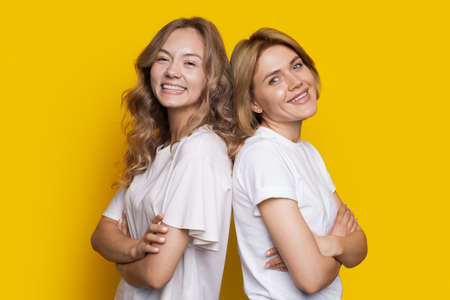Portrait of two laughing women in white t-shirts standing back to back isolated over yellow background with arms crossed. Closeup portrait.の写真素材