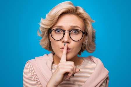 Surprised blonde haired woman makes silence gesture presses index finger over lips isolated over blue background. Shhh sign.の写真素材