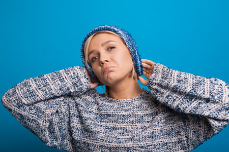 Closeup portrait of sad blonde haired woman with sincere emotions touching head wearing blue warm sweater and blue knit hat isolated over blue background.の写真素材