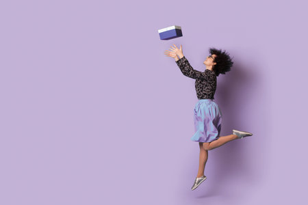 Full length body size photo of jumping caucasian curly haired woman stretching to receive gift box isolated over purple background with copy space.の写真素材