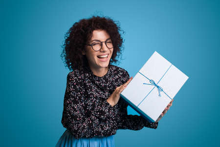 Portrait of happy curly haired girl wearing eyeglasses receiving gift box wondering whats inside isolated over blue background. Birthday presentの写真素材