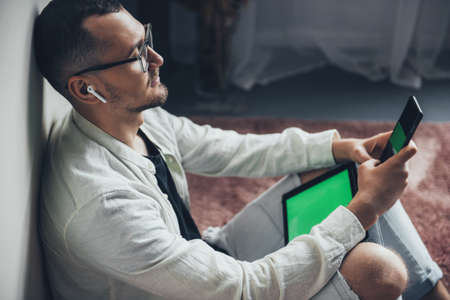 Side view of an businessman sitting on floor leaning from wallat home using digital tablet and phone...