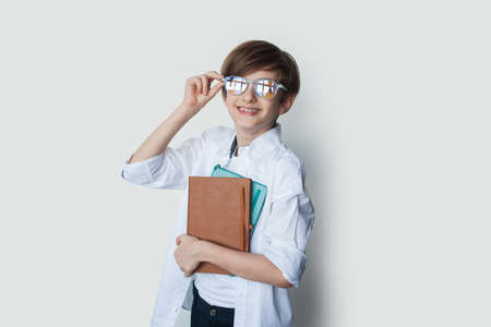 Portrait of a caucasian boy in white shirt holding a pile of copy books who stands smiling and holds his glasses, isolated on white backgroundの写真素材