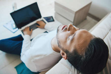 Caucasian man sleeping and laying on floor leaning his head from sofa after hard working and video conferencing.の写真素材