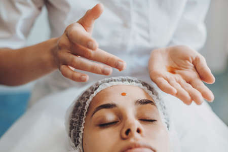 Close up portrait of hands of beautician dripping nourishing oil on forehead and facial skin of young woman in spa salon. Front view. Facial skincare. Beauty treatment. Rejuvenation treatment.の写真素材