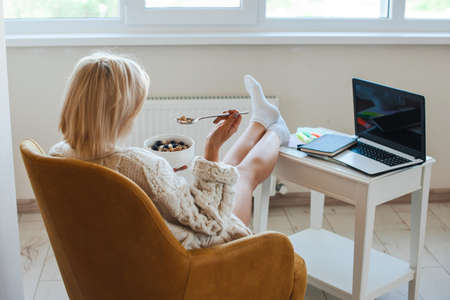 Back rear view. Woman working from home on laptop while eating the meal with muesli. Morning time....