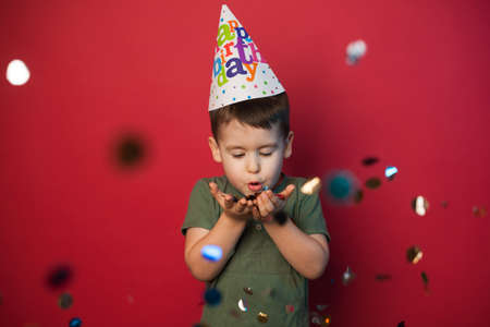 Caucasian boy in a festive cap blowing confetti to camera on a red background. The concept of a fun...