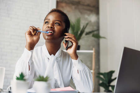 African business woman sitting by the table and talking on phone in office. Business technology. Mobile communication. Online communication. Talking people.の写真素材