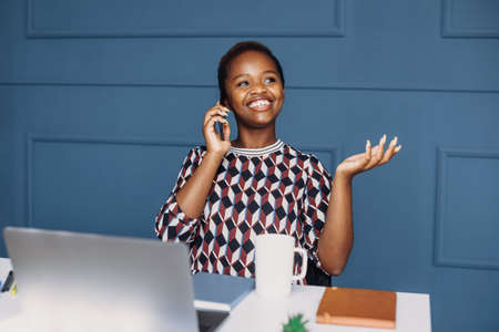 African-american business woman talking on mobile phone while working with her laptop in the office. Mobile technology. Millennial lifestyle. Using telephone. Business woman.の写真素材