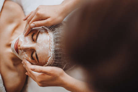 Beautician hands putting some white mask on client face in the spa salon. Top view. Beauty skin. Facial skin treatment.の写真素材