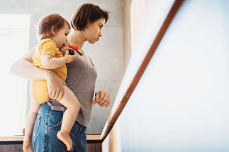 Multitasking mum holding her young baby while standing at the kitchen making food. Mother baby standing home. Family protection. Baby care.の写真素材