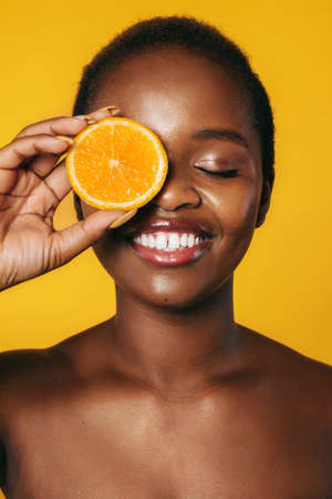 Closeup portrait of a black woman covering eye with orange slice smiling with closed eyes isolated...