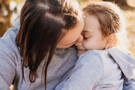 Close-up portrait of a mother kissing her daughter, whom she holds in her arms outside in the park....