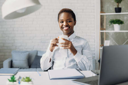 Indoor portrait of pleased african american woman looking at camera while posing in office during coffee break. Businesswoman portrait. Stylish american black portrait. Business woman. Afro american girl.の写真素材