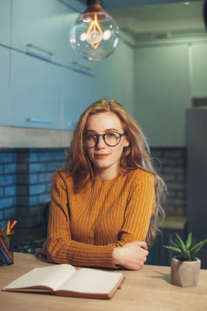 Tired redheaded woman student sitting at a table with textbooks looking at camera preparing for...