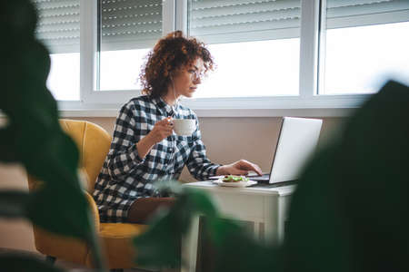 Side view of a curly business woman sitting indoos using laptop computer listening music with...