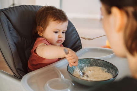 Baby girl sitting in a high chair trying to eat alone porridge with spoon. Mom feeding baby. Holding hand.の写真素材