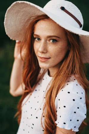 Close-up portrait of redheaded woman with freckled skin and long hair, wearing hat, posing outdoor. Beauty fashion model. Beauty skin female face. Elegant style. Hair care. Young woman skin care.の写真素材
