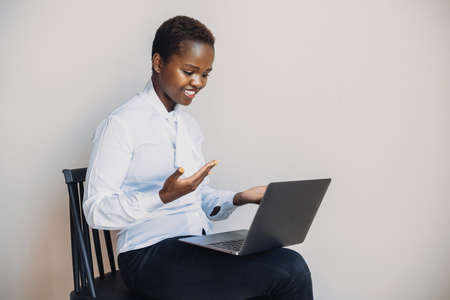 African woman sitting on a chair on white wall and working on a laptop via the internet. Beautiful...