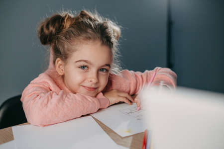 Portrait of a schoolgirl sitting at table and drawing with colourful pencils at home looking at camera and smiling. Creative idea. Smiling happy child. Enjoying preschooler development.の写真素材