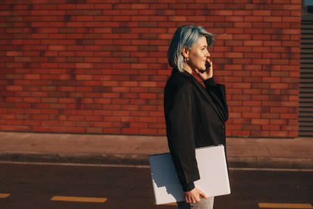 Side view of a businesswoman with short blue hair in suit having phone call and looking away while walking the street. Mobile communication. Street style. Online communication. Side view. Mobile call.の写真素材