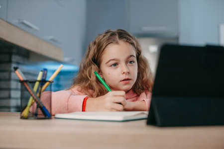 Concentrated girl listening to online lesson looking at tablet screen and taking notes. Online...