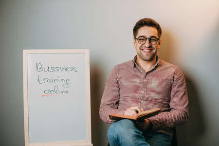 Teacher with eyeglasses holding a book in his hand sitting at home on a chair next to a white board...