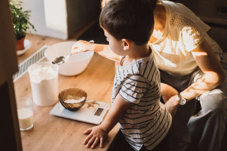 Boy helping his mother make cake dough by adding flour with a spoon. Cooking together at home kitchen.の写真素材