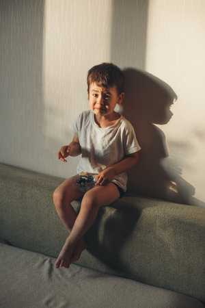 Boy wrinkling after eating a blueberry from the bowl he holds in his hands sitting on the couch. Facial expression. Healthy meal.の写真素材