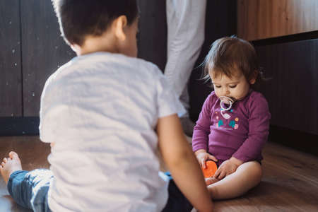Two little kids are sitting on the floor and having fun, the baby sister has a pacifier in her mouth. Family care. Baby care.の写真素材