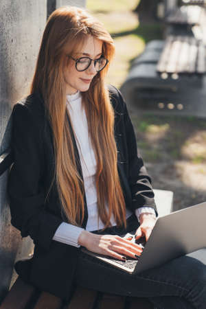 Caucasian freelancer copywriter typing on laptop, working online sitting on bench in the public park. Modern communication. Online education.の写真素材