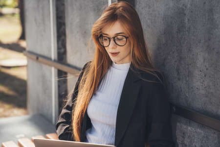 Beautiful young ginger redheaded woman in eyeglasses sitting on bench using laptop in the summer...