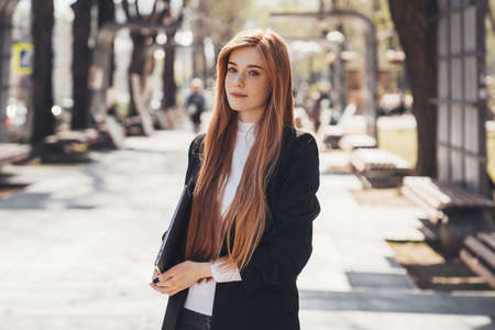 Business woman standing in the park holding a laptop in her arms waiting for her girlfriend to go to...