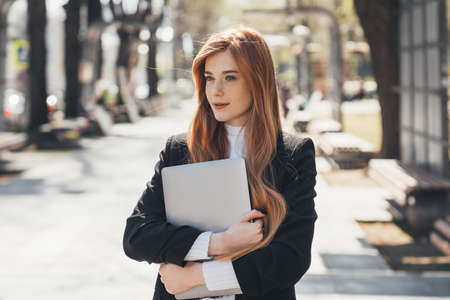 Beautiful business woman suit, with a laptop in her hands, walking outdoors in the park. Happy businesswoman. Woman studying online outdoors. Freelancer working in the park.の写真素材