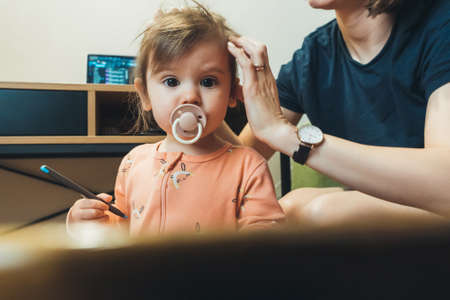 Curious baby girl looking with interest at camera, mother grooming baby hair with hand. Family care....