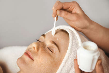 Close up portrait woman with closed eyes receiving facial rejuvenation procedure with cream in spa...