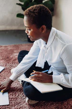 Young student girl writing ideas into book, sitting on the floor, using laptop computer. American portrait. Education internet technology.の写真素材