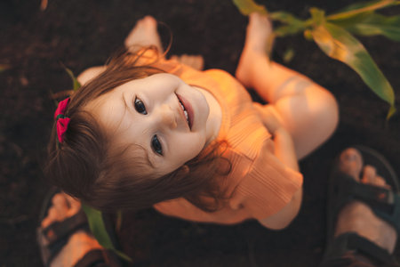 Baby girl sitting in green cornfield on sunny summer day, looking up at camera. Family exploring nature together. Country environment.の写真素材