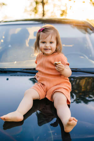 Front view of a little baby girl sitting on a hood of a car parked on a hill. Summer vacation fun. Beautiful girl portrait.の写真素材