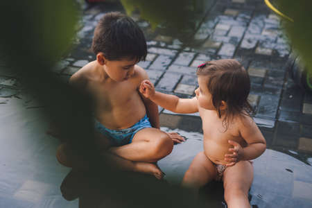 Two cute kids sitting in the yard of their house and playing with water. Spring, summer garden. Summer nature. Summer vacation fun.の写真素材