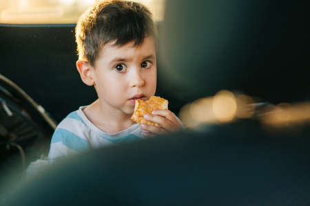 Portrait of a boy sitting in car and eating a piece of meat pie during the road trip. Summer...