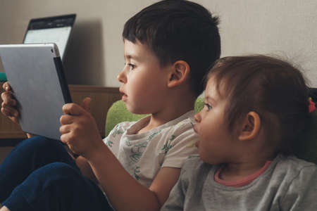 Two little kids playing games on a computer tablet together, sitting on sofa. Children and devices...