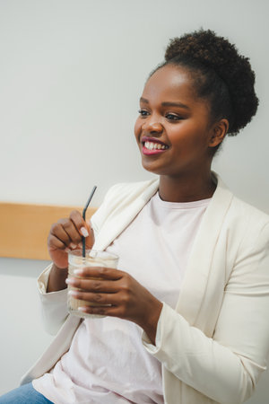 Beautiful afro woman drinking iced coffee latte using a straw indoors in a cafe. Refreshment after a...