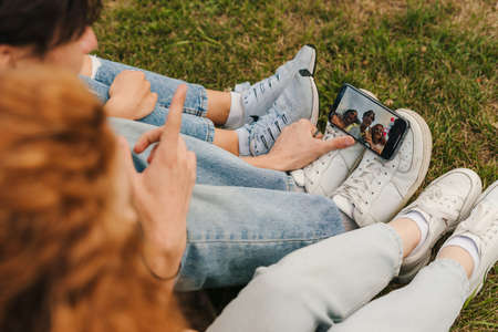 Three teenagers sitting on the grass in the park holding the phone on their shoes while making a live video to make their lives public to their followers. Young influencer vlogger. Modern communication, social media and gadgets.の写真素材
