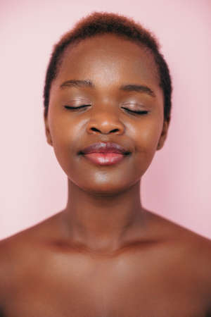 Front view of an African American woman smiling with closed eyes posing on pink background representing spa service. Attractive beautiful girl. Young woman face portrait. Facial beauty.の写真素材
