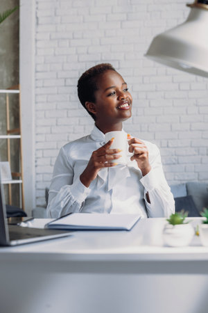 Portrait afro-american businesswoman looking away sitting at office desk drinking coffee while dreaming about weekend vacation. Business portrait. Successful multiethnic girl. Smiling girl.の写真素材
