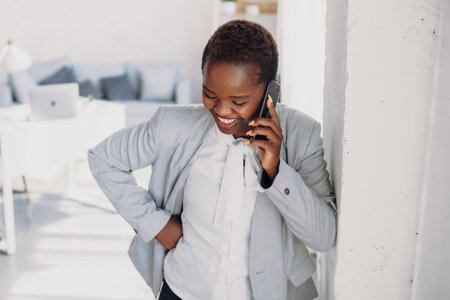 Young African American businesswoman standing in the modern office, and talking on mobile phone with the client. Modern technology. Smiling employee using cellphone. Closeup happy woman face emotionの写真素材
