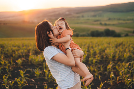 Young mother kissing her baby daughter in the green cornfield on summer day at sunset. Motherhood child care concept. Summer relaxation concept. Summer nature.の写真素材