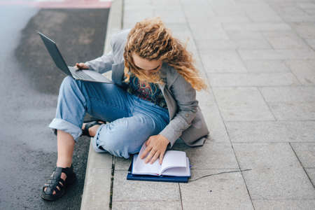 Female student in jacket studying with laptop and notepad sitting outdoors in city street preparing...